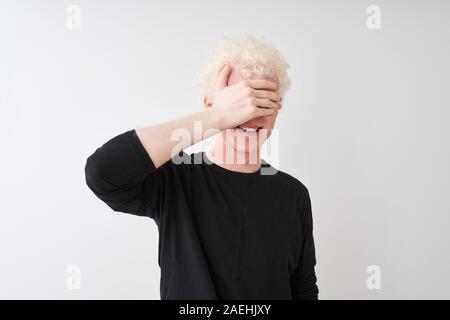 Junge albino blonder Mann in schwarzen T-Shirt stand über isoliert weißer Hintergrund zu lächeln und zu lachen mit der Hand auf das Gesicht, die Augen für Überraschung Stockfoto
