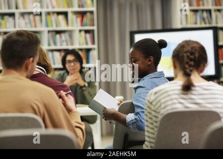 Afrikanischer junge Frau lächelnd und ein Buch lesen und diskutieren Sie mit Ihren Kollegen während der Präsentation in der Bibliothek Stockfoto