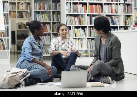 Multiethnische Gruppe von Studenten, die auf dem Boden sitzend mit Laptop und ihre Hausaufgaben besprechen im Team in der Bibliothek Stockfoto