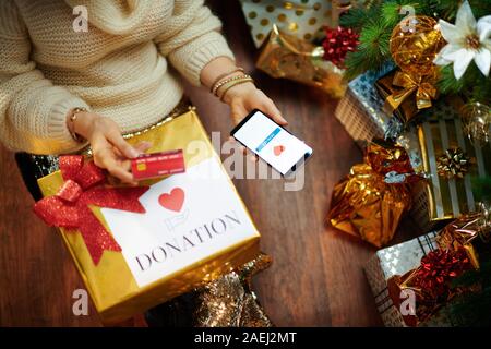 Stilvolle Hausfrau in Gold Pailletten Rock und weißen Pullover mit Kreditkarte eine Spende via Smartphone Anwendung unter dem geschmückten Weihnachtsbaum ne Stockfoto