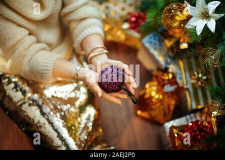 Nahaufnahme auf elegante mittleren Alter Hausfrau in Gold Pailletten Rock und weißen Pullover unter dem geschmückten Weihnachtsbaum in der Nähe des heutigen Boxen angezeigt lila Christus Stockfoto