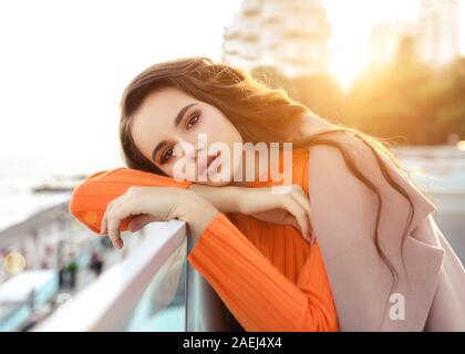 Schöne junge Mädchen mit dunklen Haaren steht in der Nähe des Meeres in einem beigen Mantel und lächelt. Sonnenuntergang auf der Sieheuntere. Stockfoto