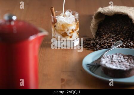 Milch in ein Glas mit kaltem Kaffee brühen, crushed Eis und einer Zimtstange umgeben von einen Sack mit Kaffeebohnen, Kuchen und eine französische Presse gießen Stockfoto