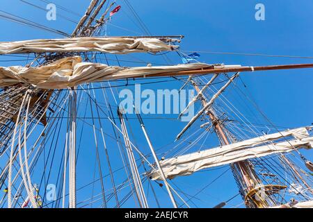 Hölzerne Masten eines alten Segelschiffs oder eines großen Schiffes mit aufgerollten weißen Segeln, Fahnen und Seilen gegen einen sattblauen Himmel. Stockfoto