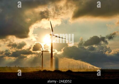 Zwei Windkraftanlagen und die Bewässerung von Erntegut Wasserpistole oder Wassersprühstrahl bei Sonnenuntergang im Gegenlicht gegen ein orangefarbener dramatische Wolkenhimmel und helle Sonne Stockfoto