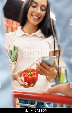 Die täglichen Einkäufe. Paar in den Supermarkt mit Warenkorb zusammen mit Paprika surfen Smartphone lächelt Fröhlich close-up Stockfoto