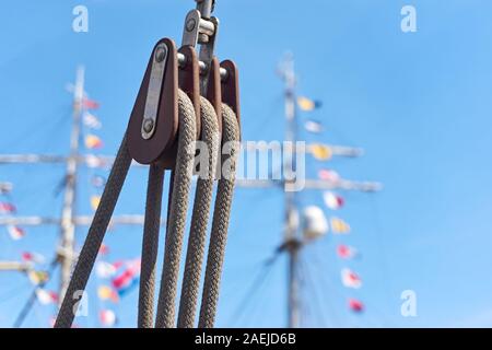 Nahaufnahme einer modernen Seilrolle mit Seilen gegen einen blauen Himmel und Masten mit farbigen Fahnen und selektivem Fokus. Stockfoto