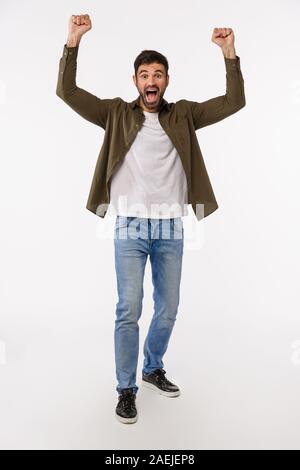 Erfolg, Leistung und Sieg Konzept. Vertikale in voller Länge Schuß froh und glücklich Siegen junger bärtiger Mann in Jeans und Mantel, heben die Hände bis in Stockfoto