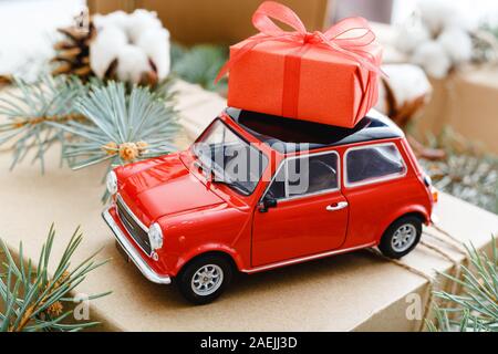 Rote Spielzeugauto mit einem weihnachtlichen roter Geschenkbox auf dem Dach. Weihnachten Hintergrund mit Baumwolle, tannenzweigen, Tannen Zapfen. Weihnachten, Feier Stockfoto