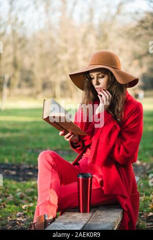 In der Nähe der schöne Brünette junge Frau insgesamt rot Look im Herbst Park. Ein Mädchen in einem braunen Hut mit breiter Krempe ist leidenschaftlich über das Lesen ein Buch heraus Stockfoto