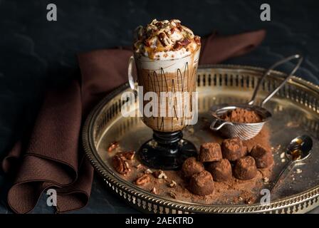 Ein dekoratives Glas Cappuccino mit Sahne, Pekannüsse, Karamell Nieselregen und Kakao besprüht mit Schokolade Trüffel serviert. Stockfoto