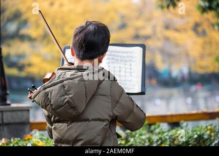 Chengdu, Provinz Sichuan, China - Dez 8, 2019: Junge Junge spielt Geige in der Straße von Jinjiang Fluss in Jinli ZhongLu Straße im Herbst mit gelben Blätter auf Ginkgo Bäume im Hintergrund. Stockfoto