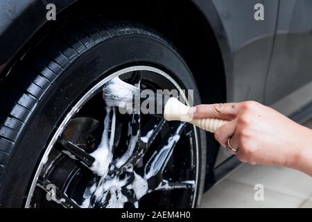 Ein männlicher Arbeitnehmer wäscht ein schwarzes Auto mit einer speziellen Bürste für Gegossene Räder und scheuert sich die Oberfläche zu glänzen in einem Fahrzeug mit Werkstatt. Auto Service in Stockfoto