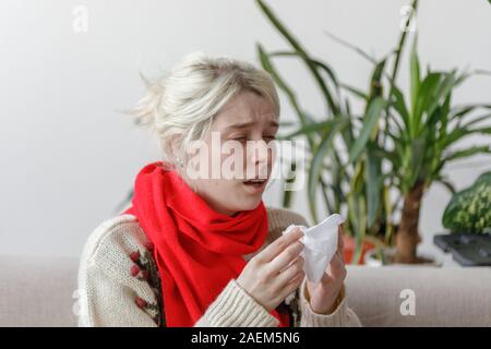 Mädchen in einem Pullover niest, während er auf dem Sofa. Der Patient erkältet, Krankheitsgefühl und Niesen in einer Papierserviette. Stockfoto