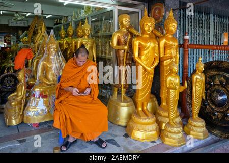 Ein buddhistischer Mönch sitzt zwischen Buddha Statuen in einem Geschäft für Buddhistische Artefakte, versessen Peering auf seinem Mobiltelefon; Bamrung Muang Rd., Bangkok, Thailand Stockfoto