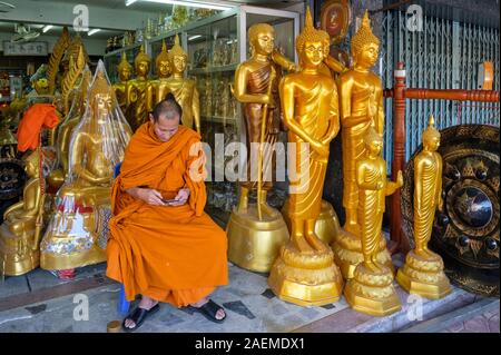 Ein buddhistischer Mönch sitzt zwischen Buddha Statuen in einem Geschäft für Buddhistische Artefakte, versessen Peering auf seinem Mobiltelefon; Bamrung Muang Rd., Bangkok, Thailand Stockfoto