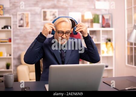 Ältere Mann in seinem 60s mit Kopfhörern auf dem Kopf Musik hören und arbeiten auf einem modernen Laptop Stockfoto