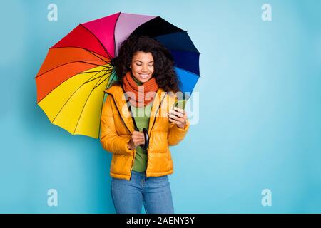 Foto der schönen dunklen Haut Dame halten bunten Regenschirm lesen e-mail Telefon modernen Blogger tragen Mantel jeans Grünen pullover schal isoliert Blau Stockfoto