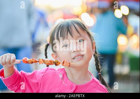Wenig asiatisches Mädchen zu Fuß auf der Straße von Bangkok, Grill kaufen und essen Essen am Abend Zeit Stockfoto