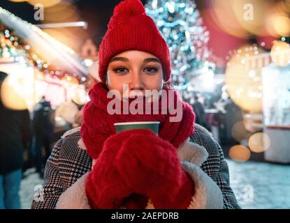 Junge schöne Frau, gekleidet in roten Schal und Red Hat Holding in Rot Handschuhe heißer Glühwein auf europäischer Weihnachtsmarkt und lächelnd. Atmosphäre Stockfoto
