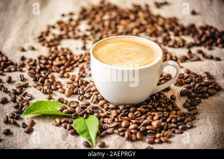 Eine Tasse Kaffee mit Schaum und grüne Blätter auf die verstreuten Körner auf die Leinwand. Duftende belebende Getränk aus frisch gerösteten Kaffeebohnen. Stockfoto