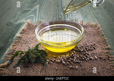 Gießen Hanföl in eine Glasschüssel und Hanf Samen in einem Löffel aus Holz, auf einem grünen Hanf-blatt Hintergrund, CBD Hanföl Stockfoto