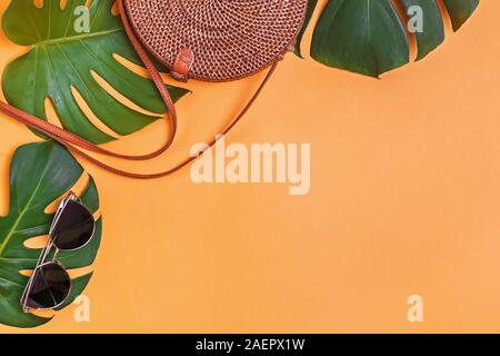 Runde Rattan Tasche und Sonnenbrille liegen auf einem monstera treibt auf gelbem Hintergrund Stockfoto