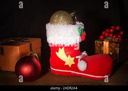 Red Santa boot mit Weihnachten Geschenke, Lollypop und Dekoration Kugeln auf dunklem Hintergrund. Happy holidays Komposition. Stockfoto