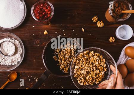 Woman's Hand hinzufügen gehackte Walnüsse in geschmolzene Schokolade in Kochtopf unter Zutaten für Backwaren Schokoladenkuchen Kuchen auf rustikalen Hintergrund, Ansicht von oben. Stockfoto