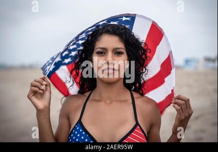 Dunkelhaarige Latina hält Amerikanische Flagge über dem Kopf beim Tragen der Sterne und Streifen Bikini am Strand am 4. Juli 2009 Stockfoto