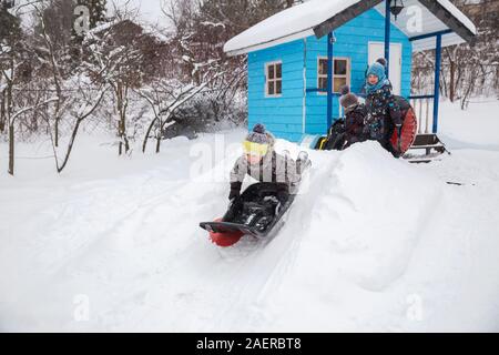 Kinder Rodeln im Winter im Hof. Drei kleine Kinder in warmen Overalls spielen draußen auf frostigen Wintertag. Drei Brüder nehmen Schlittenfahrt wiederum her Stockfoto