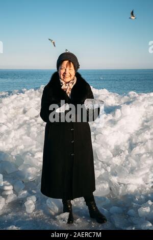 Senior lächelnd schöne Frau in schwarzen Mantel holding Brocken von Meereis vor der hügeligen Eis Stockfoto