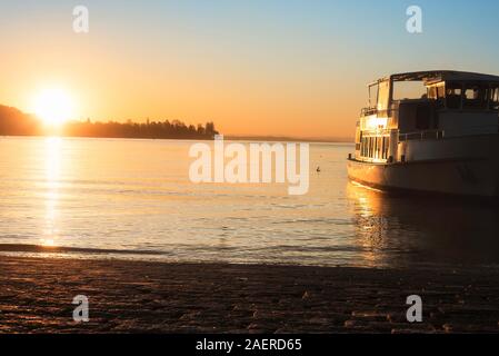 Sonnenaufgang über Wasser und ein Boot am Bodensee, in Konstanz, Deutschland. Bodensee Seeufer und einem Schiff Kreuzfahrt in Richtung Sonnenaufgang. Wasser Navigation. Stockfoto