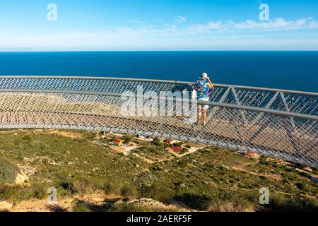 Frau mittleren Alters tragen ein Sonnenhut auf den Blick von der Santa Pola Skywalk Aussichtsplattform auf den Klippen oberhalb Partida Bancal de la Arena, EIN Stockfoto