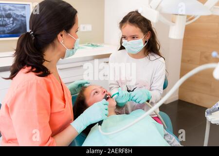 Adorable Kinder Zahnarzt in der zahnärztlichen Chirurgie mit zwei hübschen Mädchen zeigen Ihnen zahnmedizinische Check-up, ein Mädchen ist Assistent spielen Stockfoto