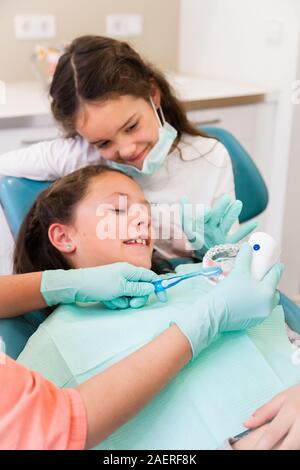 Adorable Kinder Zahnarzt in der zahnärztlichen Chirurgie mit zwei hübschen Mädchen zeigen Ihnen zahnmedizinische Check-up, ein Mädchen ist Assistent spielen Stockfoto