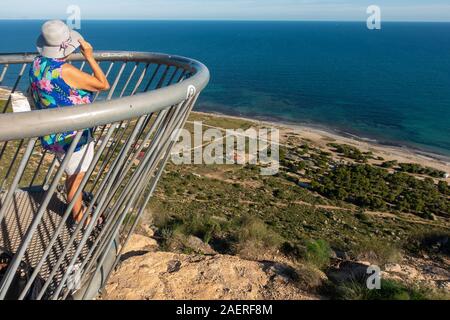 Frau mittleren Alters tragen ein Sonnenhut auf den Blick von der Santa Pola Skywalk Aussichtsplattform auf den Klippen oberhalb Partida Bancal de la Arena, EIN Stockfoto