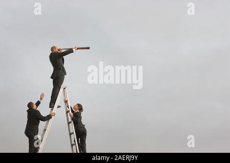 Ein Geschäftsmann stehen auf der obersten Sprosse einer hohen trittleiter schaut durch ein Teleskop als männliche und weibliche Kollegin Aufstieg auf der Leiter. Stockfoto