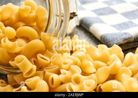 Pipe Rigate pasta Rollen aus einem offenen Glas storage jar. Im Hintergrund eine verschwommene blau kariertem Tuch. Stockfoto