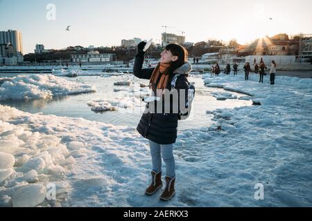 Odessa, Ukraine - Februar, 16 2017: junge lächelnde Frau, die auf gefrorenen Küste des Schwarzen Meeres mit Eis holding Stück Eis bei Sonnenuntergang fällt. Stockfoto