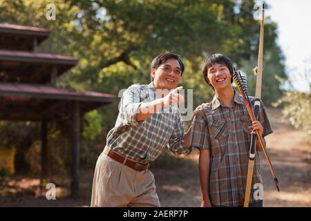 Lächelnd Mitte der erwachsene Mann Lehre sein halbwüchsiger Sohn wie ein Pfeil und Bogen zu benutzen. Stockfoto