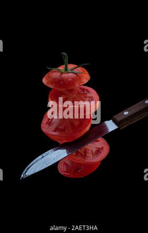 Levitation von frisch geschnittenen Tomaten. Ein Messer schneidet Tomaten in der Luft. 5 fliegen Tomatenscheiben auf einem schwarzen Hintergrund. Stockfoto