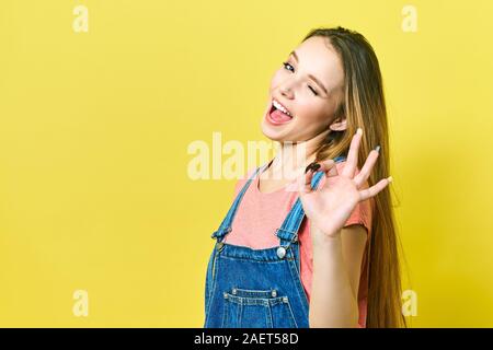 Lustige und fröhliche Mädchen zeigt okay unterzeichnen und zwinkert Kamera auf gelbem Hintergrund. Stockfoto