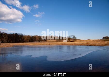 Schöne Landschaft mit See fast bedeckt mit dünnem Eis. Winter oder Frühling Szene an einem sonnigen Tag mit blauem Himmel, weißen Wölkchen und einen Wald in Stockfoto