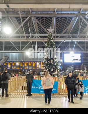 Die geschmückten Weihnachtsbaum in der bahnhofshalle Hauptbahnhof von Glasgow, Schottland, mit Passagieren zu betrachten, während andere auf ihre Züge bewegen. Anzeigetafel im Hintergrund. Stockfoto