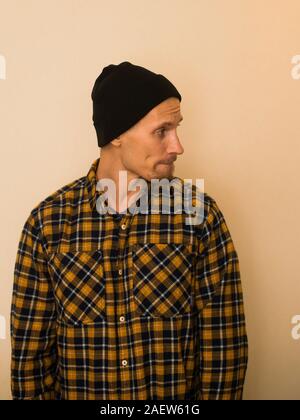 Ein junger Kerl in einem Plaid Shirt stellt gegen eine beige Wand. Modell in der alltäglichen Kleidung. Stilvolle junger Mann. Stockfoto