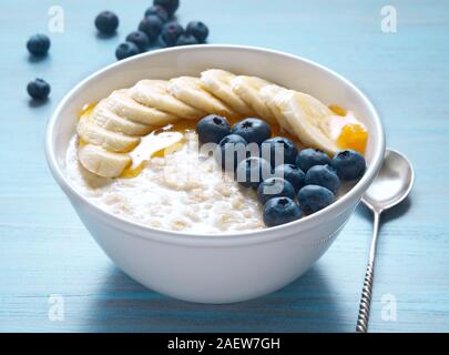 Haferflocken mit Bananen, Blaubeeren, Chia Samen, Marmelade, Honig auf einem blauen Hintergrund aus Holz. Gesundes Frühstück. Von der Seite. Stockfoto