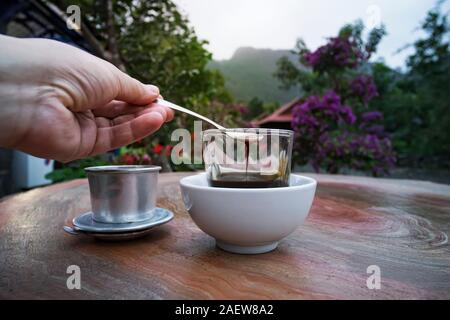 Vietnamesischen Kaffee auf dem Tisch vor dem Hintergrund der schönen Vietnamesischen Natur in einem Straßencafe. Der Mann rührt den Kaffee mit einem Löffel. Teelöffel Stockfoto