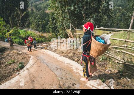 Mädchen aus dem Stamm der Roten Dzao, mit einem großen Weidenkorb hinter, von Kindern umgeben. TA PHIN, Lao Cai, VIETNAM - 12. Januar 2019 Stockfoto