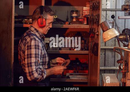 Ältere Arbeitnehmer tragen Overalls und Kopfhörer Auswählen einer Festplatte für Schleifer beim Stehen in der Werkstatt Stockfoto